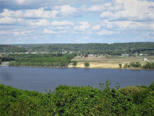 A scenic view of a river bordered by lush greenery and fields under a partly cloudy sky.