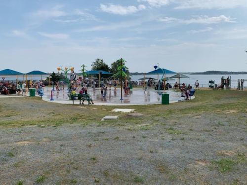 A vibrant splash pad with children playing, surrounded by green grass and a view of the water in the background.