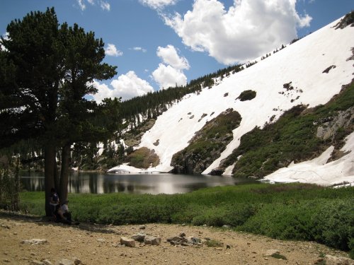 A serene landscape featuring a lake surrounded by snow-capped mountains and lush greenery under a partly cloudy sky.
