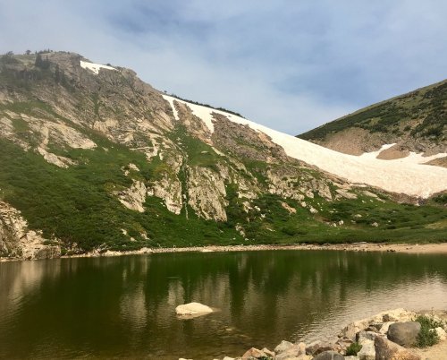 A serene mountain landscape featuring a green hillside, a snow-capped peak, and a calm lake reflecting the scenery.