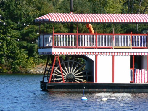 A colorful riverboat with a large paddle wheel, docked on a calm lake surrounded by greenery.