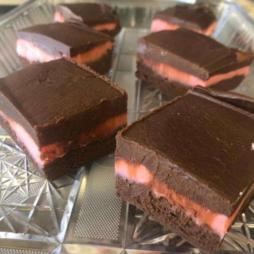 A close-up of chocolate-covered layered squares with a pink filling, arranged on a clear tray.