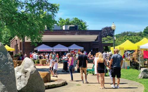 A lively outdoor festival with people walking, colorful tents, and greenery under a clear blue sky.
