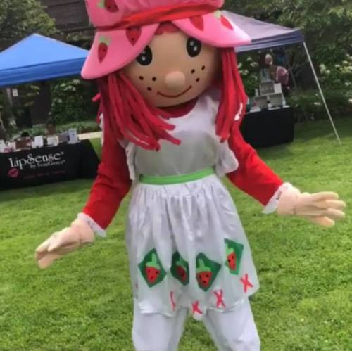 A person in a colorful strawberry-themed mascot costume stands outdoors, smiling in a park setting.