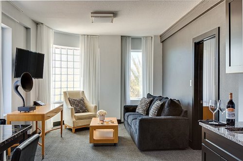 A modern hotel room featuring a sofa, armchair, TV, and a small table with wine and glasses. Bright natural light.