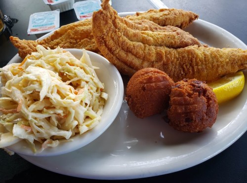 A plate of fried catfish fillets, coleslaw, and two cornbread muffins, garnished with a lemon wedge.