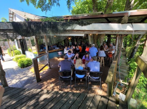 A lively outdoor dining area with people seated at tables under trees, enjoying a sunny day at a restaurant.