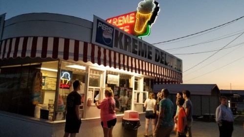 A busy ice cream stand at dusk, with a neon sign and a line of customers ordering treats.