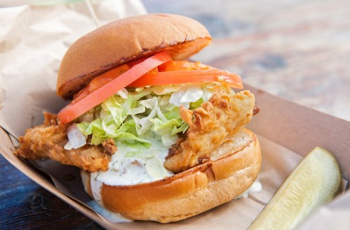 A crispy fried chicken sandwich with lettuce, tomato, and sauce, served with a pickle on the side.