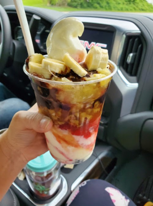 A hand holding a large dessert cup filled with layers of ice cream, bananas, chocolate syrup, and strawberries.