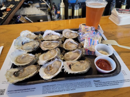 A platter of fresh oysters on ice, accompanied by cocktail sauce, crackers, and a pint of beer at a bar.