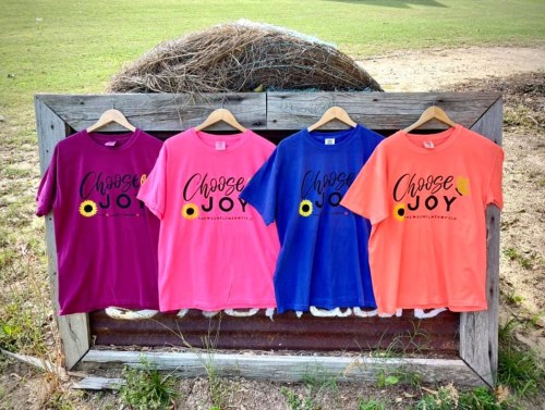 Four colorful t-shirts hanging on a wooden display, each featuring the phrase "Choose Joy" with a sunflower design.