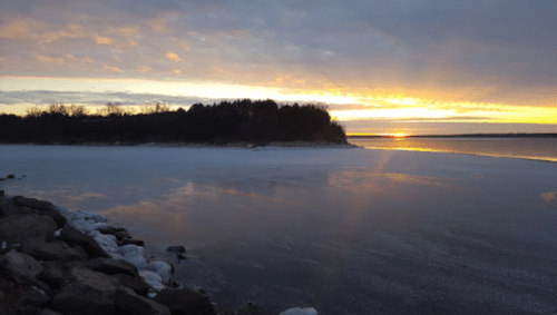 Sunset over a frozen lake, with rocky shoreline and trees silhouetted against a colorful sky.