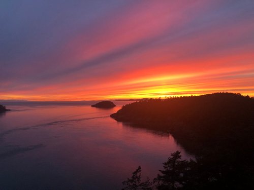 A vibrant sunset over calm waters, with silhouettes of islands and trees against a colorful sky.