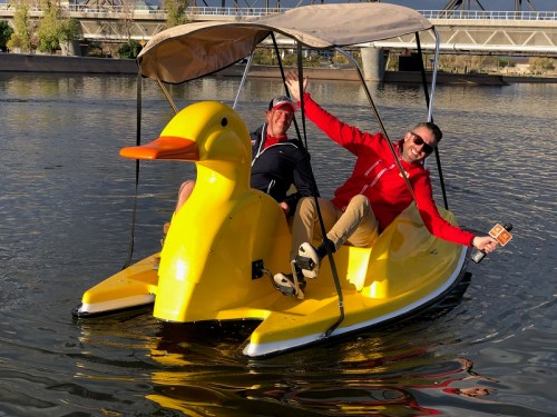 Two men joyfully pose on a large yellow duck-shaped paddle boat on a calm waterway.