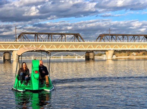 A couple enjoys a pedal boat ride on a calm lake, with a bridge and cloudy sky in the background.