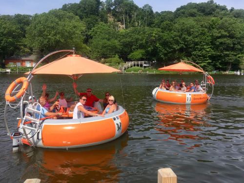 Two orange floating boats on a lake with people enjoying themselves, surrounded by green trees and a sunny sky.