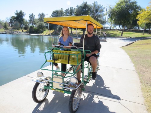 A man and woman smile while riding a green pedal boat by a lake on a sunny day. Trees line the shore in the background.