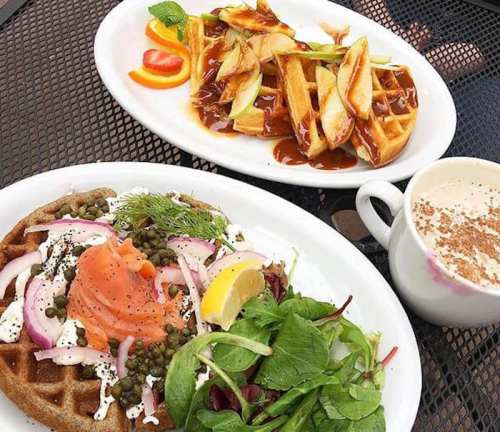 A plate with smoked salmon on a waffle, salad, and a cup of coffee, alongside another plate of waffles with toppings.