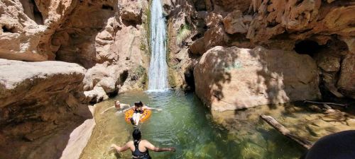 A group of people enjoying a natural pool with a waterfall, surrounded by rocky cliffs and greenery.