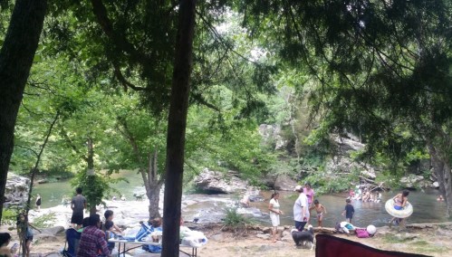 A lively river scene with people swimming, relaxing, and enjoying nature among trees and rocks.