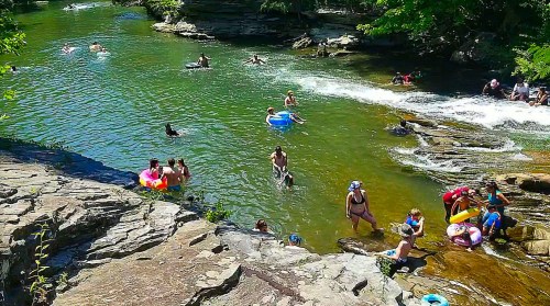 A lively river scene with people swimming and floating on tubes, surrounded by rocks and greenery on a sunny day.