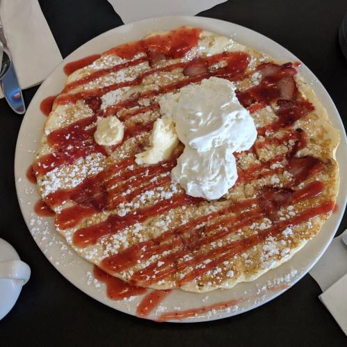 A large pancake topped with whipped cream, strawberry sauce, and powdered sugar on a white plate.