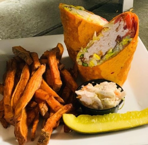 A plate featuring a cut burrito filled with vegetables, alongside sweet potato fries, coleslaw, and a pickle.