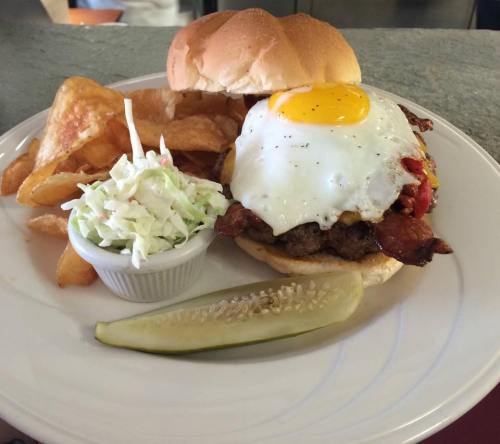 A burger topped with a fried egg, served with potato chips, coleslaw, and a pickle on a white plate.