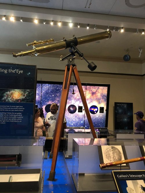 A vintage telescope on a wooden tripod, displayed in a museum with a starry background and informational panels.