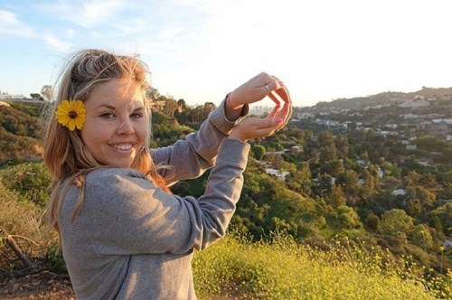 A smiling woman with a flower in her hair holds her hands in a circle, overlooking a scenic hillside at sunset.