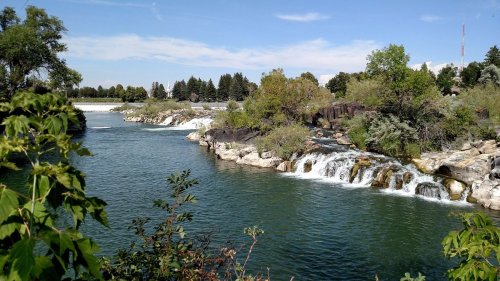 A serene river flows past rocky banks and lush greenery, with a small waterfall cascading into the water under a clear sky.