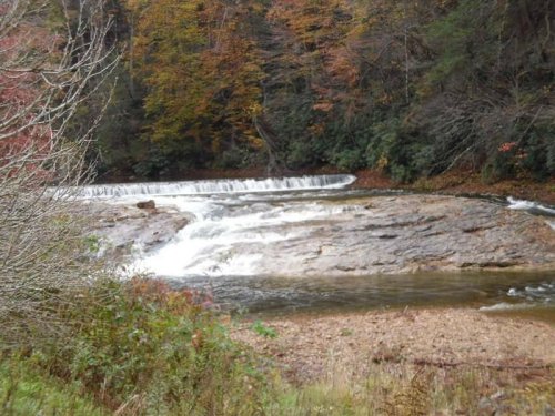 A serene waterfall cascades over rocky terrain, surrounded by autumn foliage and a peaceful riverbank.