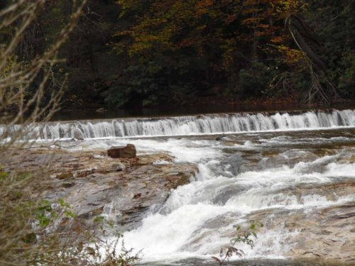 A serene river flows over rocky terrain, with gentle waterfalls and autumn foliage in the background.