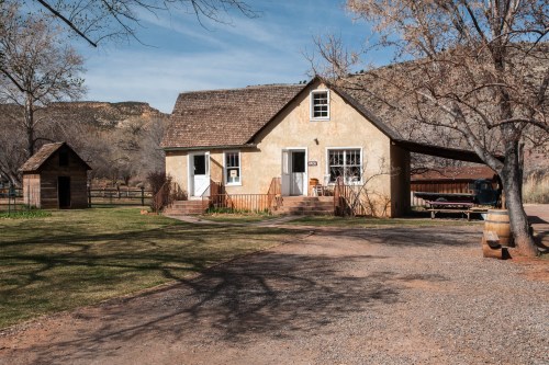A quaint yellow house with a sloped roof, surrounded by trees and a gravel path, set against a mountainous backdrop.