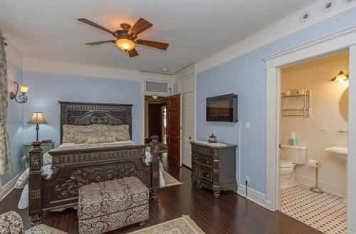 Cozy bedroom with a dark wooden bed, ceiling fan, and en-suite bathroom, featuring light blue walls and elegant decor.