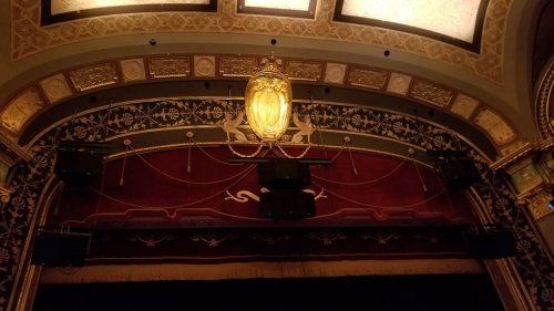 Ornate theater ceiling with decorative gold details and a central light fixture above a red velvet curtain.