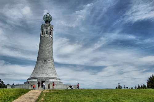 A tall stone monument with a lantern atop, surrounded by green grass and people enjoying the view under a cloudy sky.