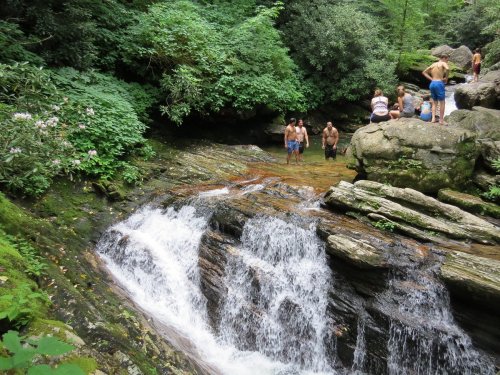 A serene waterfall flows over rocks, with people enjoying the water and lush greenery surrounding the scene.