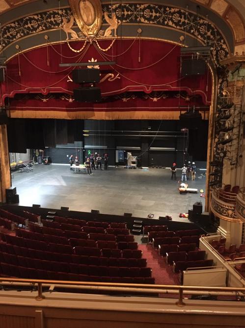 A theater stage with crew members setting up, red velvet seats visible in the foreground, and ornate decor in the background.
