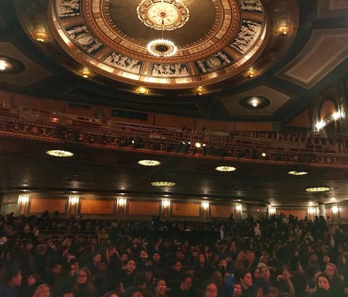 A crowded theater interior with ornate ceiling details and warm lighting, filled with an audience eagerly awaiting a performance.