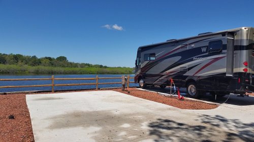 A parked RV by a serene lake, surrounded by greenery and a wooden fence under a clear blue sky.