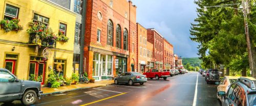 A charming street lined with colorful buildings, shops, and parked cars under a cloudy sky.