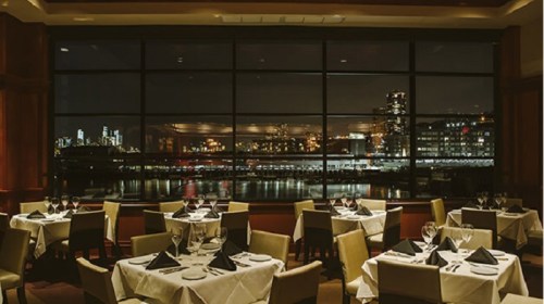 Elegant restaurant interior with tables set for dining, featuring a large window showcasing a city skyline at night.