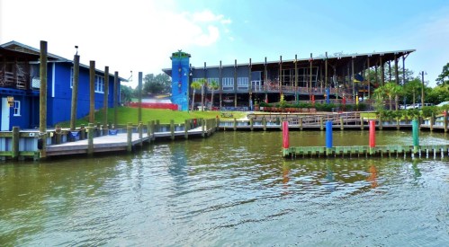 A waterfront view of a colorful building with a tower, surrounded by docks and greenery under a blue sky.