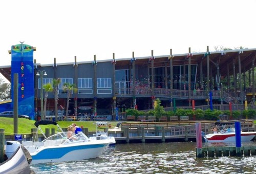 A waterfront restaurant with a large deck, boats docked nearby, and colorful flags flying in a sunny setting.