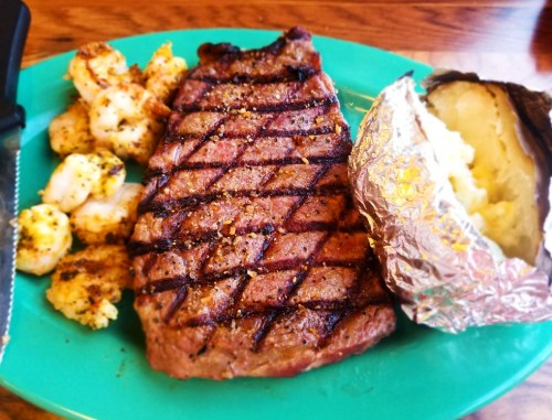 A grilled steak with crosshatch marks, shrimp, and a baked potato wrapped in foil on a green plate.
