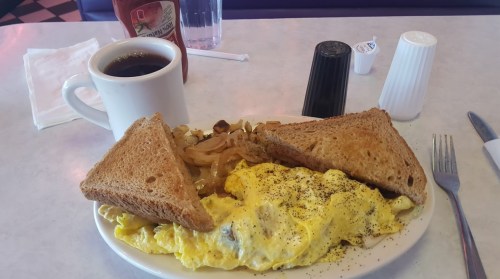 A plate with an omelette, toast, hash browns, and a cup of coffee on a diner table. Ketchup and condiments are nearby.