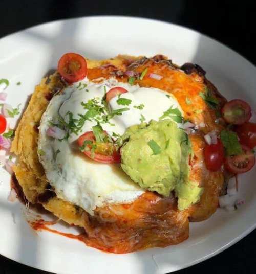 A plate of enchiladas topped with a fried egg, guacamole, cherry tomatoes, and fresh herbs.