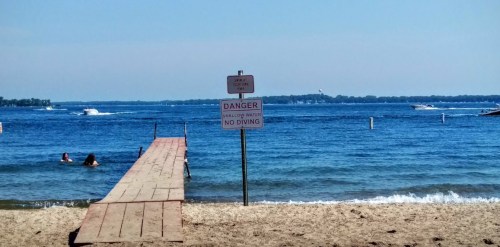 A sandy beach with a wooden dock and a warning sign about shallow water and no diving, with boats in the background.